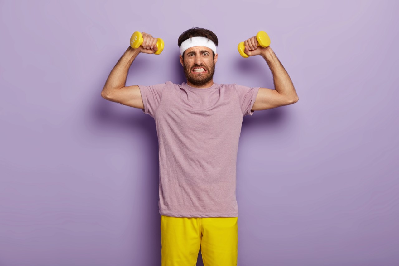 determined-young-sporty-unshaven-man-clenches-teeth-raises-muscular-arms-does-exercises-with-dumbbells-clenches-teeth_1280x853